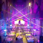 Event lighting image from back of the monastery manchester with Multi colour beams of moving lights, stage with two large screens and moving lights on truss stands with uplights making them shine in pink and blue