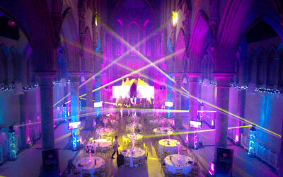 Event lighting image from back of the monastery manchester with Multi colour beams of moving lights, stage with two large screens and moving lights on truss stands with uplights making them shine in pink and blue